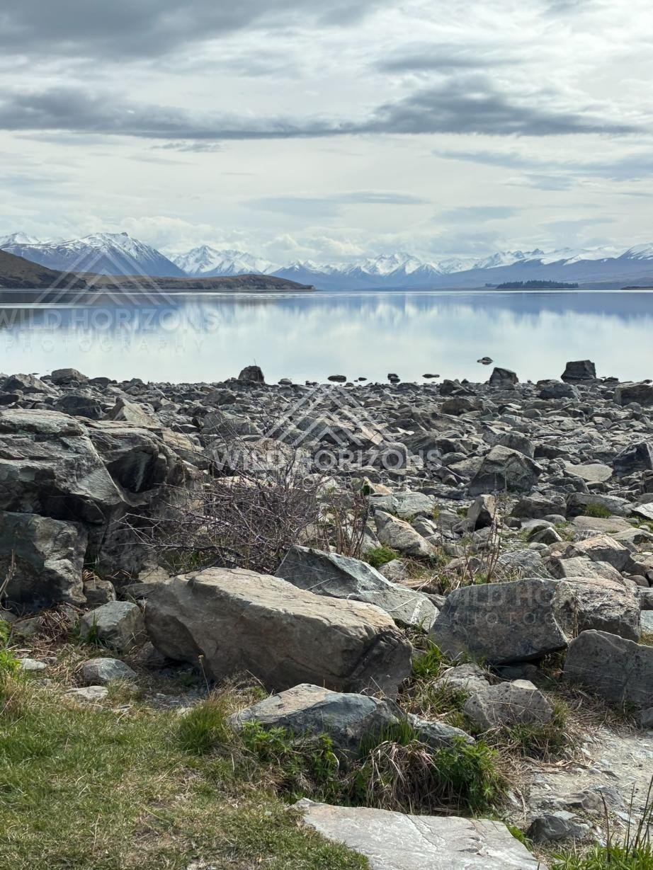 Rocky lakeshore with calm reflections beneath distant alpine ranges. Lake Tekapo, New Zealand