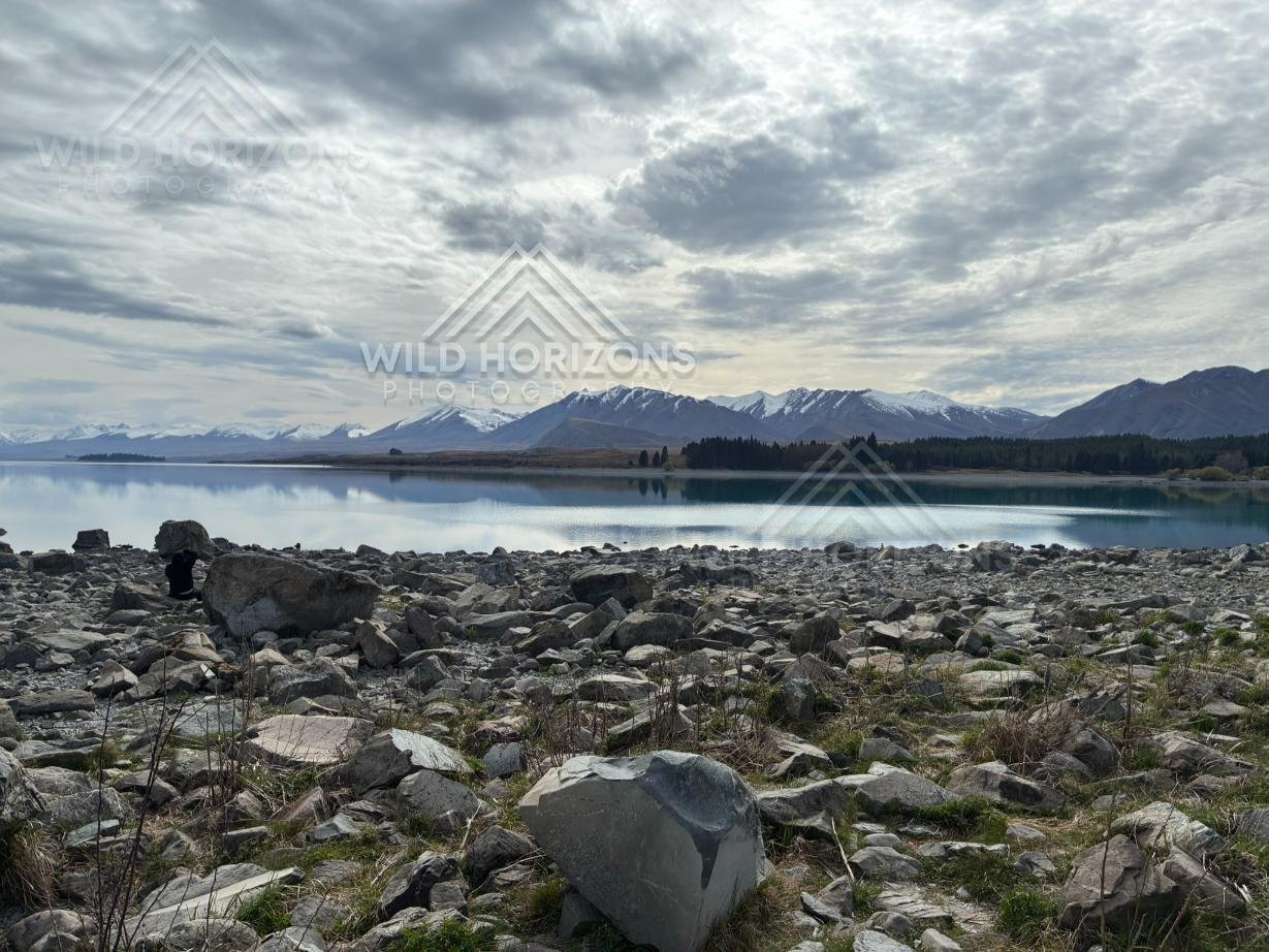 Rocky Lakeshore and Reflected Mountain Range under Overcast Skies. Lake Tekapo, New Zealand