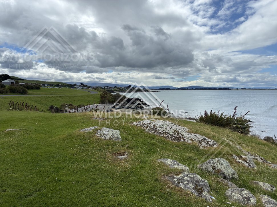 Grassy foreshore with scattered homes under dramatic cloud above calm coastal water. Riverton, New Zealand
