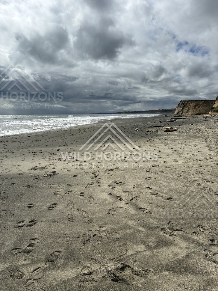 Wide black-sand beach stretching beneath heavy cloud and rolling surf. Southland coast, New Zealand