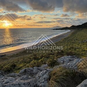 Sunset Dropping Toward the Sea Along a Long Curving Beach and Rocky Foreground. Riverton, New Zealand