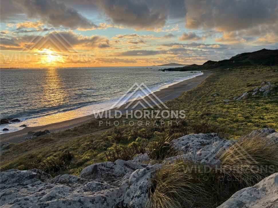 Sunset Dropping Toward the Sea Along a Long Curving Beach and Rocky Foreground. Riverton, New Zealand
