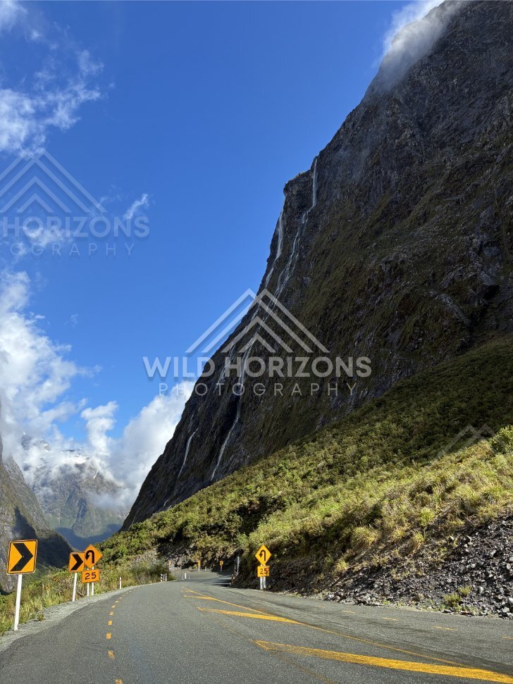 Curve Warning Signs on the Milford Road Below a Towering Waterfall, Milford Road, Fiordland National Park, New Zealand