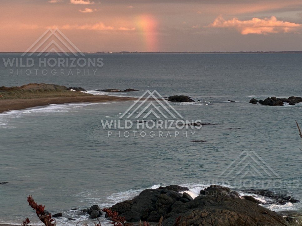 Rainbow over rocky shoreline and calm sea. Riverton, New Zealand.