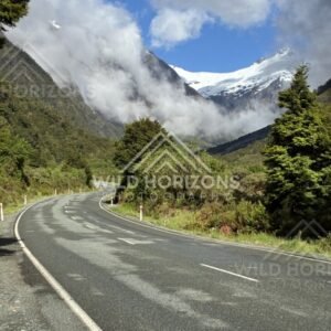 Milford Road Curve Through Forest Toward Cloud-Wrapped Mountains. Milford Road, New Zealand.