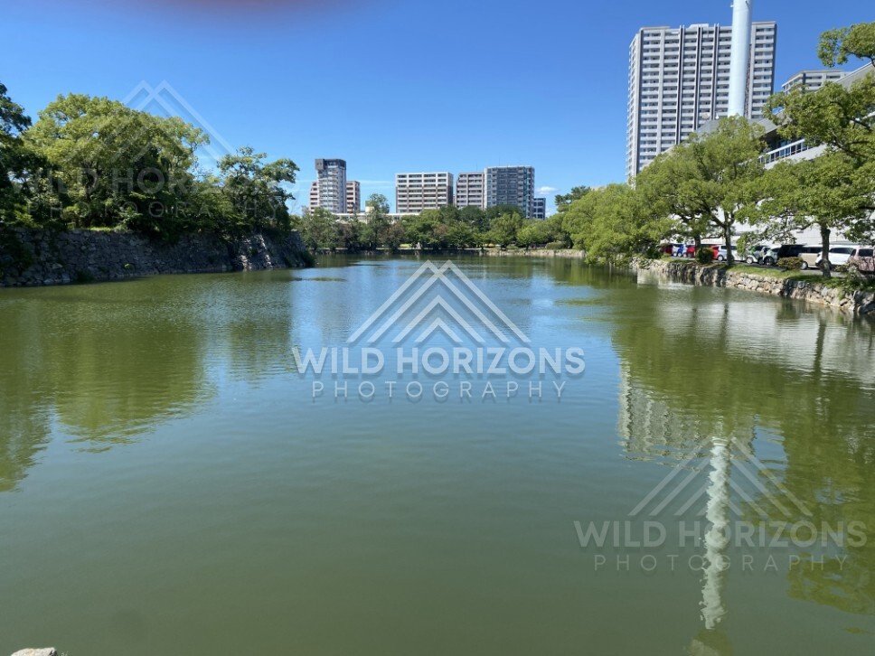 Moat waters reflecting city buildings beside a tree-lined embankment. Hiroshima, Japan.