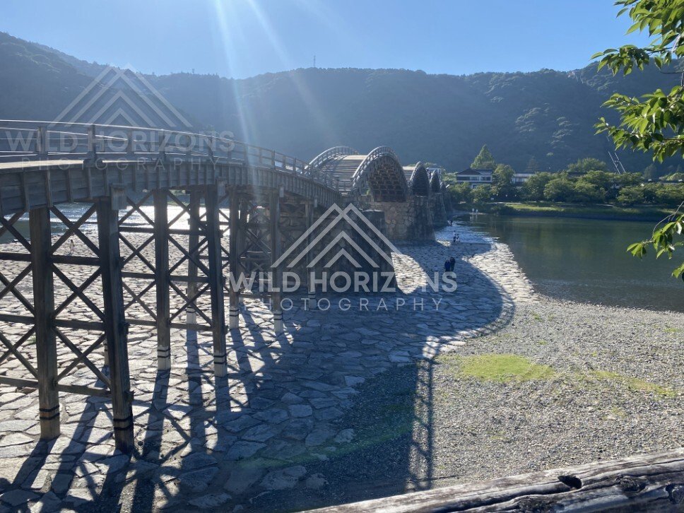 Sunlit wooden bridge curving across a dry riverbed toward forested hills. Iwakuni, Yamaguchi, Japan.