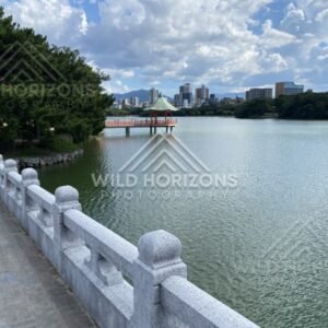 A stone-railed walkway follows the lakeshore toward a pavilion over the water. Fukuoka, Japan.