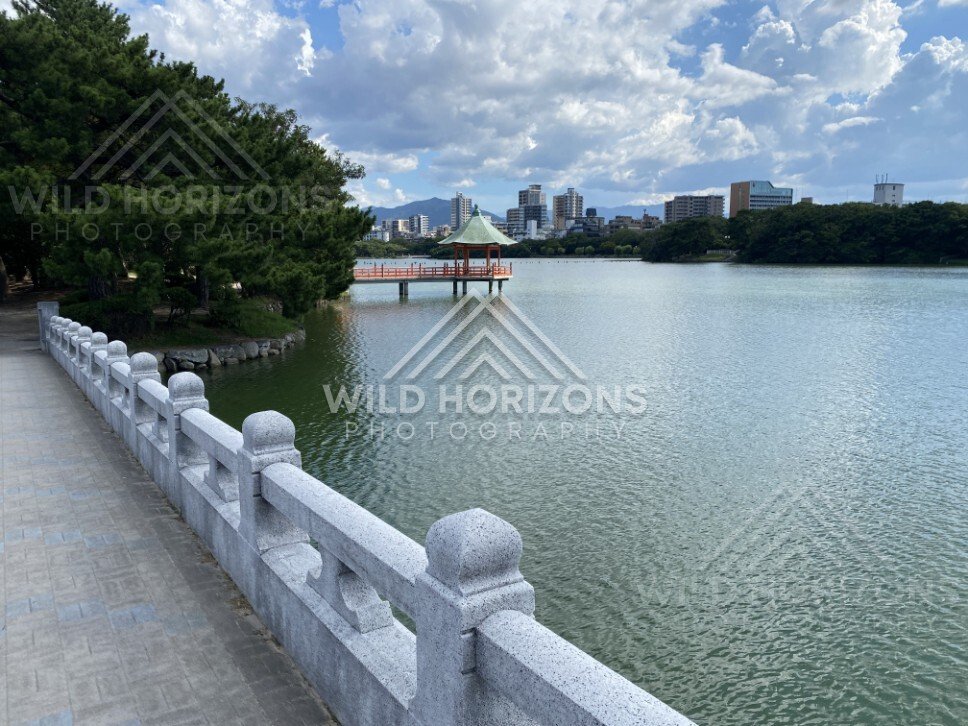 A stone-railed walkway follows the lakeshore toward a pavilion over the water. Fukuoka, Japan.