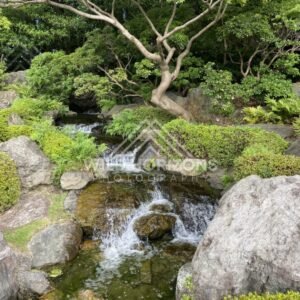 A small waterfall cascades over stones beneath leafy trees in a Japanese garden. Fukuoka, Japan.