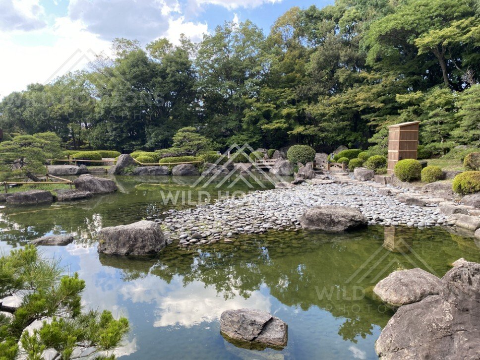 A rock-and-pebble shoreline meets a reflective pond inside a Japanese garden. Fukuoka, Japan.