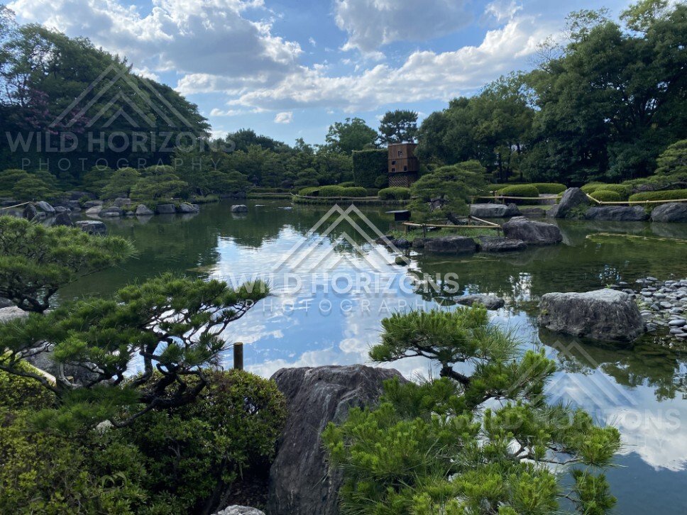 Clouds reflect in a still pond with boulders and clipped pines in the foreground. Fukuoka, Japan.