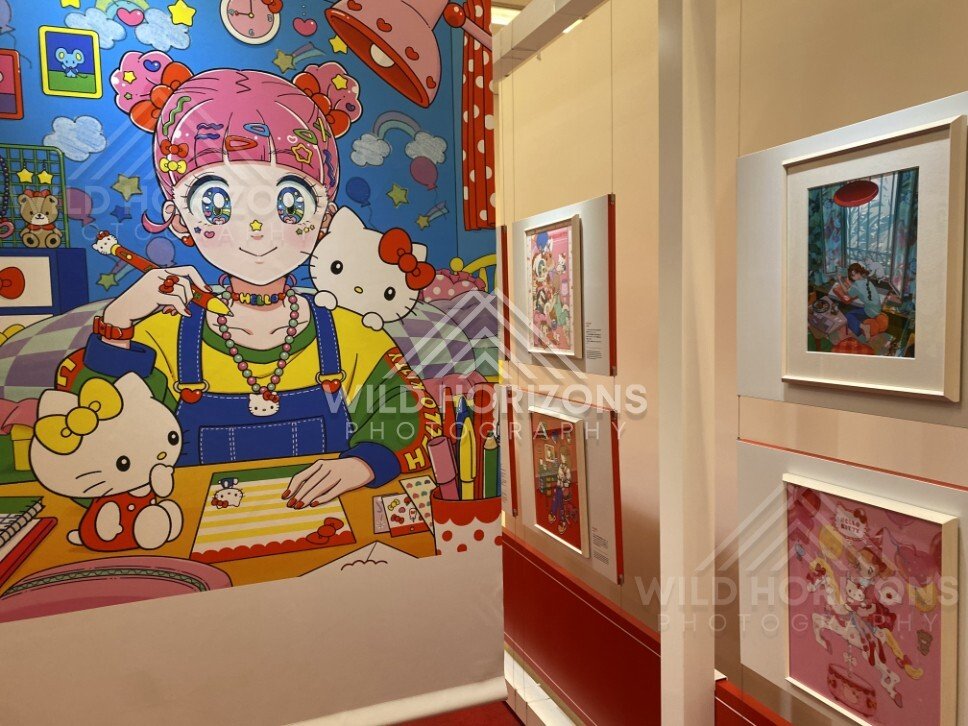 A large Hello Kitty mural stands beside framed artworks in a brightly lit gallery. Fukuoka, Japan.