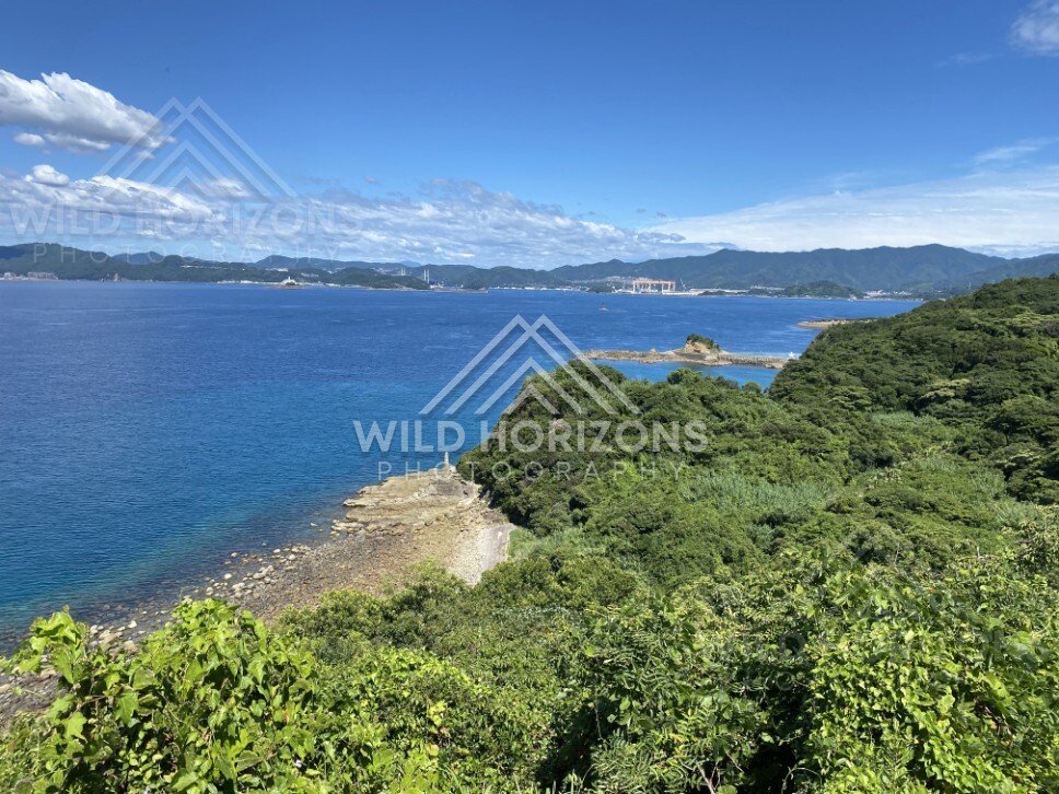 Rocky shoreline and forested peninsula overlooking clear blue coastal waters. Nagasaki, Japan.