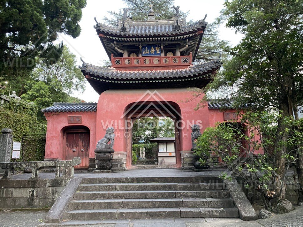 Ornate red temple entrance gate framed by trees and stone steps. Nagasaki, Japan.