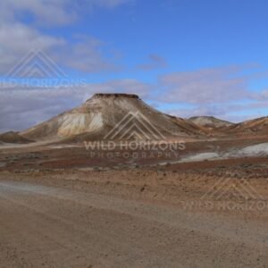 Graded outback track curves past a layered Breakaways mesa under clearing blue sky. Kanku-Breakaways Conservation Park/Coober Pedy, Australia.