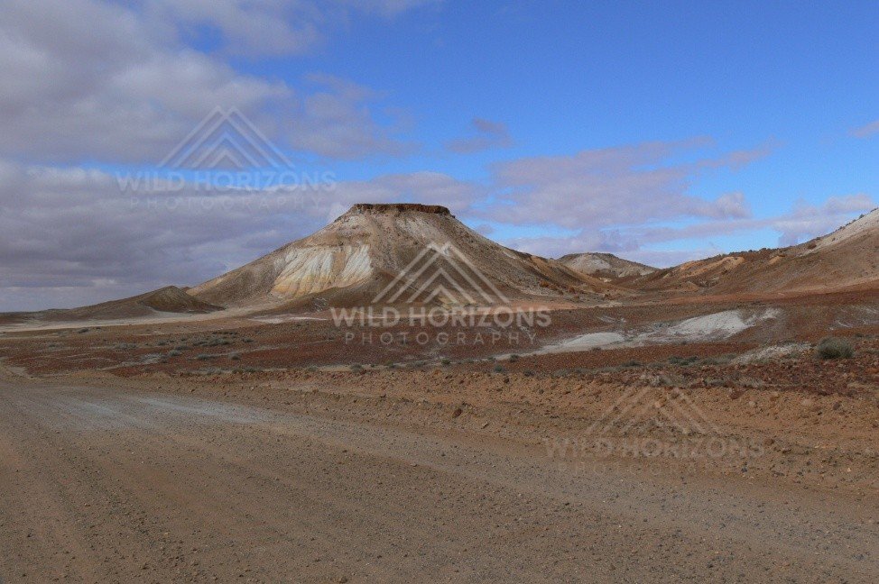 Graded outback track curves past a layered Breakaways mesa under clearing blue sky. Kanku-Breakaways Conservation Park/Coober Pedy, Australia.