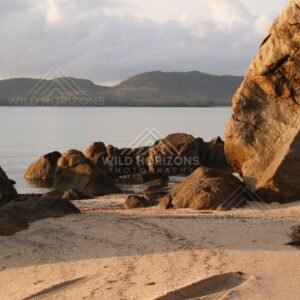 Granite boulder on a sandy beach with calm water and distant hills. Portland Roads, Australia.