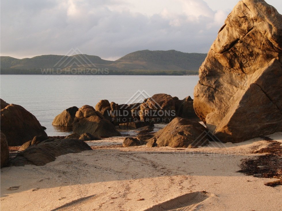 Granite boulder on a sandy beach with calm water and distant hills. Portland Roads, Australia.