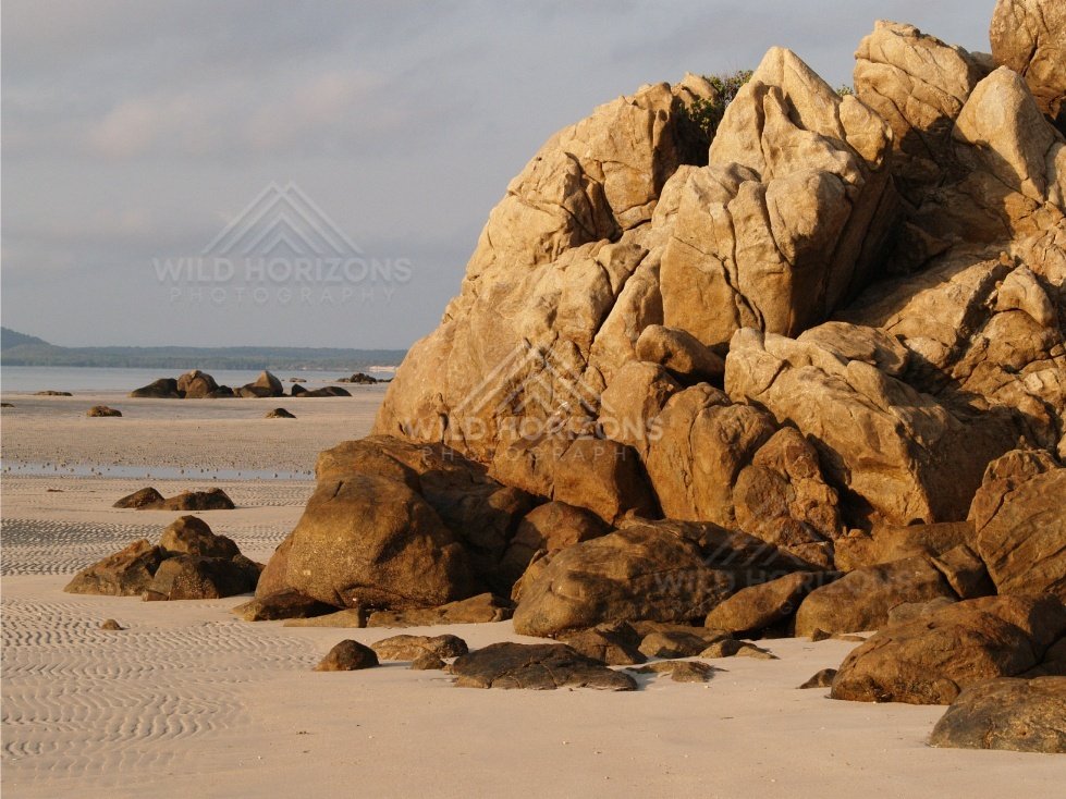 Sunlit granite boulders rising from a sandy beach at low tide. Portland Roads, Australia.