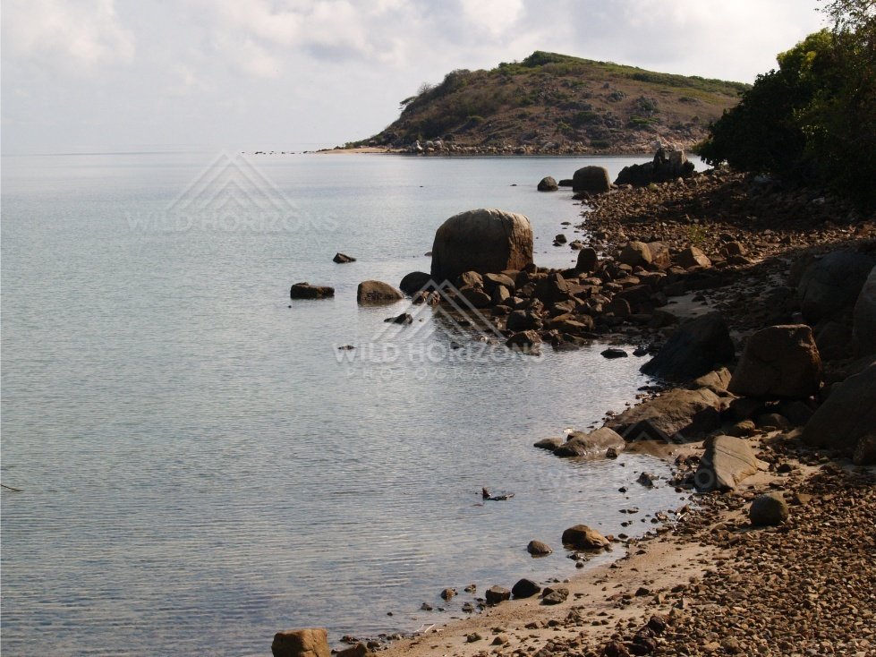 Rocky shoreline with small stones and calm water beside a forested headland. Portland Roads, Australia.
