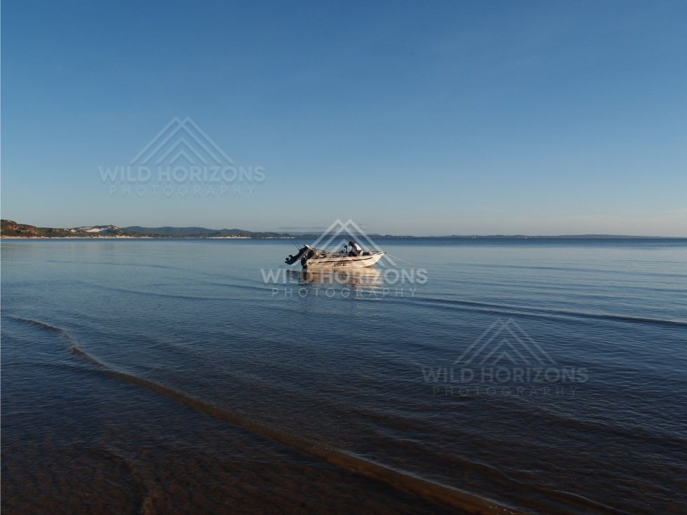 Small boat anchored on calm water with sandbars and distant coastline. Elim Beach, Australia.